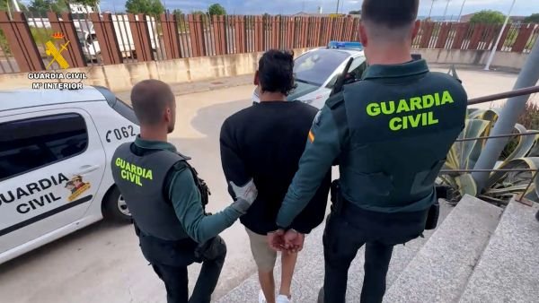 Dos agentes de la Guardia Civil escoltan por la espalda a un detenido esposado hacia los vehículos patrulla oficiales.