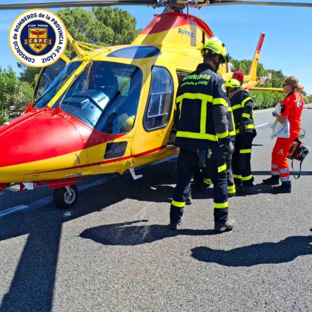 Bomberos de la provincia de Cádiz junto a personal sanitario al lado de un helicóptero amarillo y rojo de emergencias de Avincis en la AP-4.