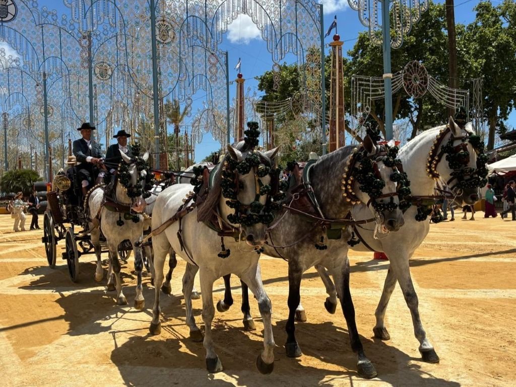 Coche de caballos tradicional tirado por cuatro ejemplares tordos desfilando por el Real de la Feria del Caballo de Jerez bajo el alumbrado y un cielo azul despejado.