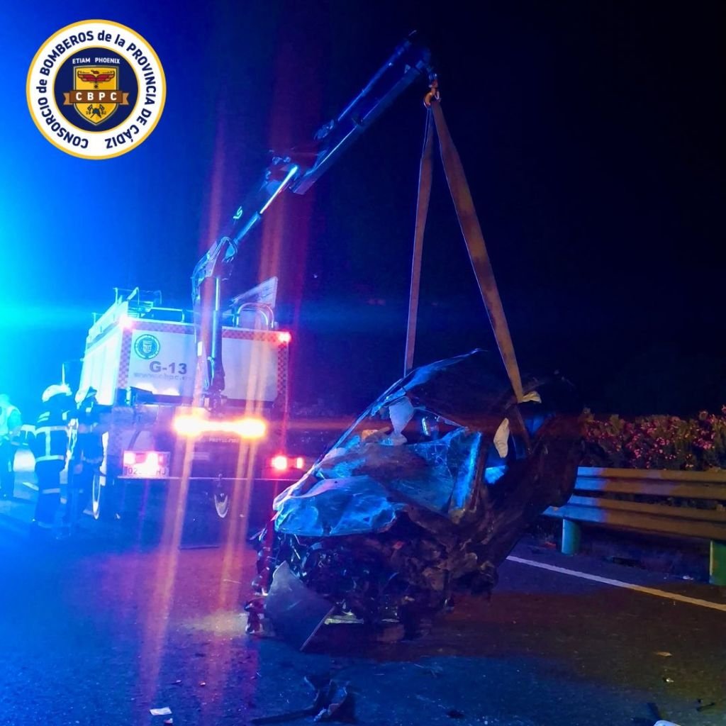 Coche siniestrado y severamente deformado suspendido en el aire por las correas de un camión grúa de los Bomberos de Cádiz durante una intervención nocturna en la carretera A-382.