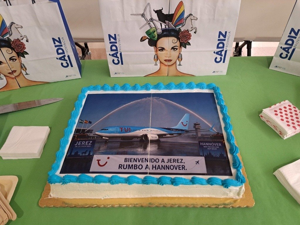 Tarta conmemorativa decorada con la imagen del avión de TUI bajo un arco de agua y bolsas promocionales con el logotipo de la Diputación de Cádiz sobre una mesa lista para la celebración.