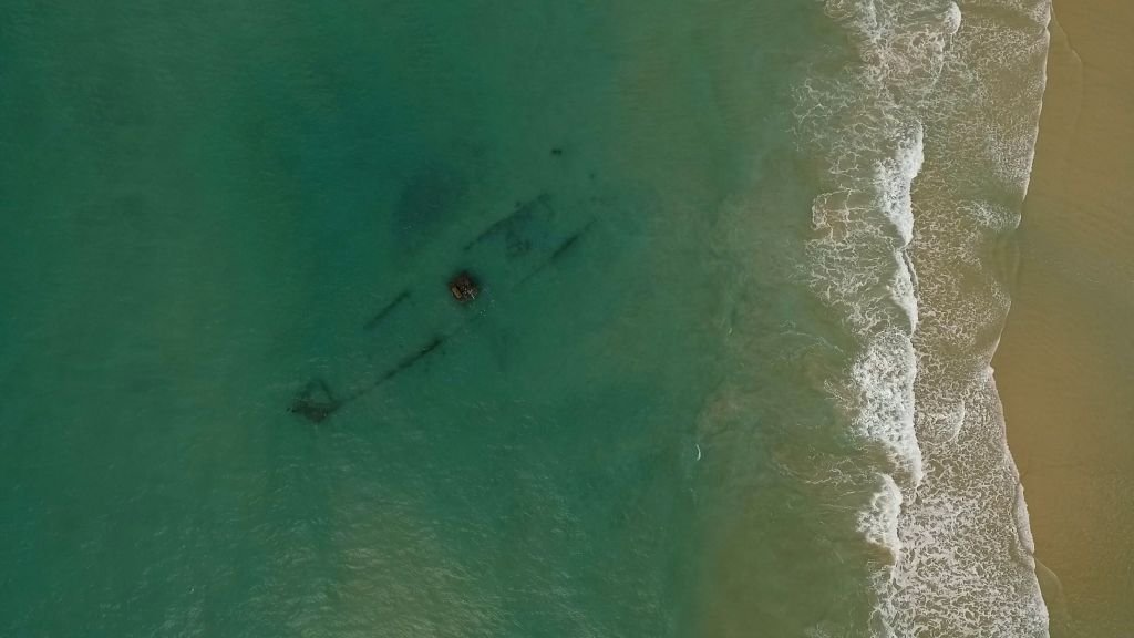 Vista aérea con dron que muestra la silueta sumergida de un antiguo barco hundido cerca de la orilla de una playa de arena dorada y aguas turquesas en Cádiz.