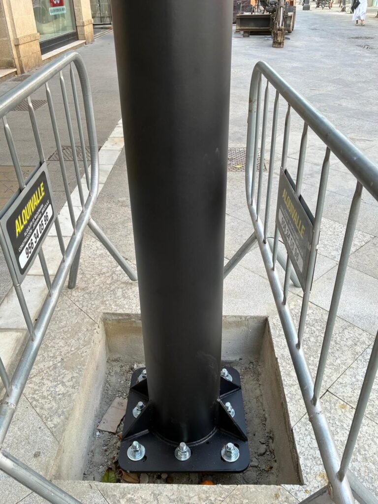 Detalle de la base de un poste negro metálico anclado firmemente con gruesos tornillos al suelo de la calle Larga de Jerez, protegido por vallas de seguridad metálicas durante las obras.