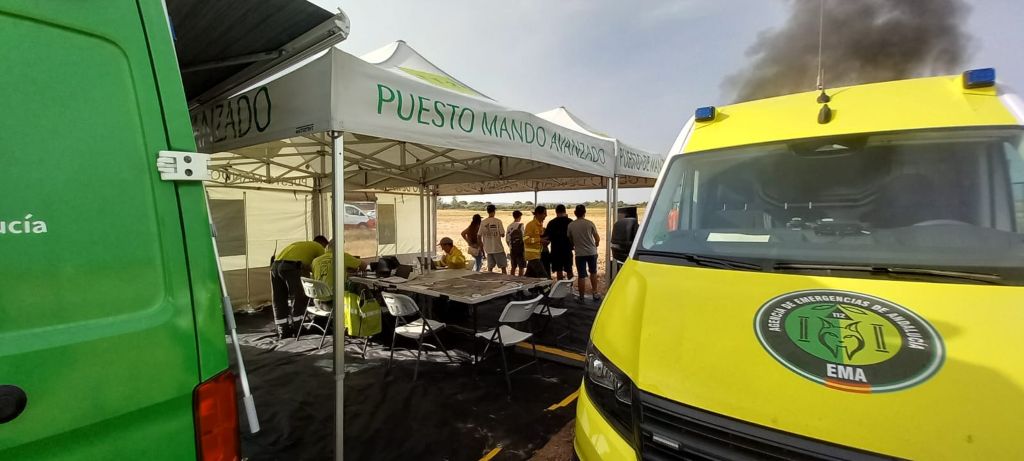 Imagen de un operativo de emergencia en campo abierto durante un simulacro. En primer plano, la parte delantera de una furgoneta de emergencia amarilla con el logotipo de EMA (Agencia de Emergencias de Andalucía 112) y una furgoneta verde a la izquierda. Detrás de ellas, una carpa blanca tipo toldo sirve de Puesto de Mando Avanzado, con personal uniformado de amarillo trabajando en mesas con mapas y ordenadores. Al fondo, un grupo de civiles espera y una columna de humo negro se eleva desde detrás de la furgoneta amarilla.