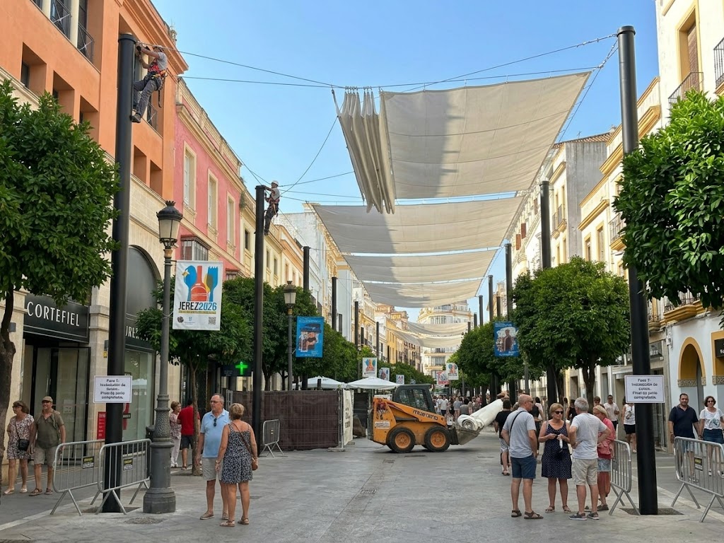 Operarios trabajando en la altura sobre báculos metálicos para fijar y desplegar las lonas blancas de los nuevos toldos de la calle Larga de Jerez, con peatones transitando por el centro comercial abierto.