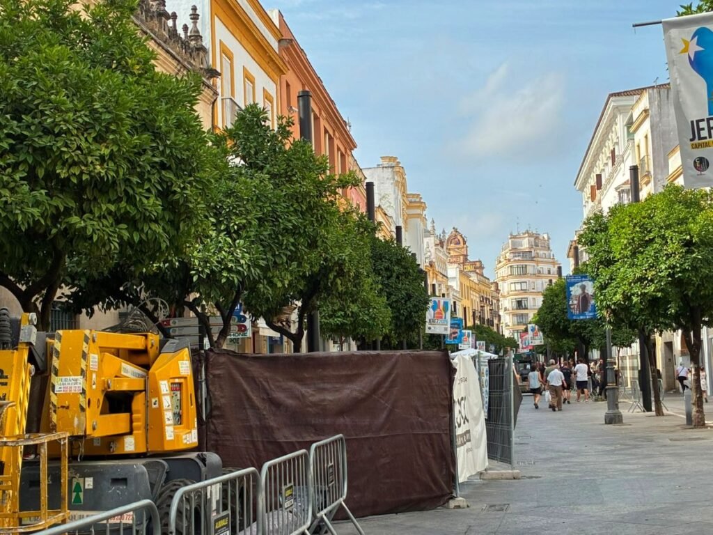 Maquinaria elevadora de color amarillo y vallas de protección junto a los naranjos de la calle Larga de Jerez durante las obras de instalación de los nuevos báculos metálicos para los toldos.