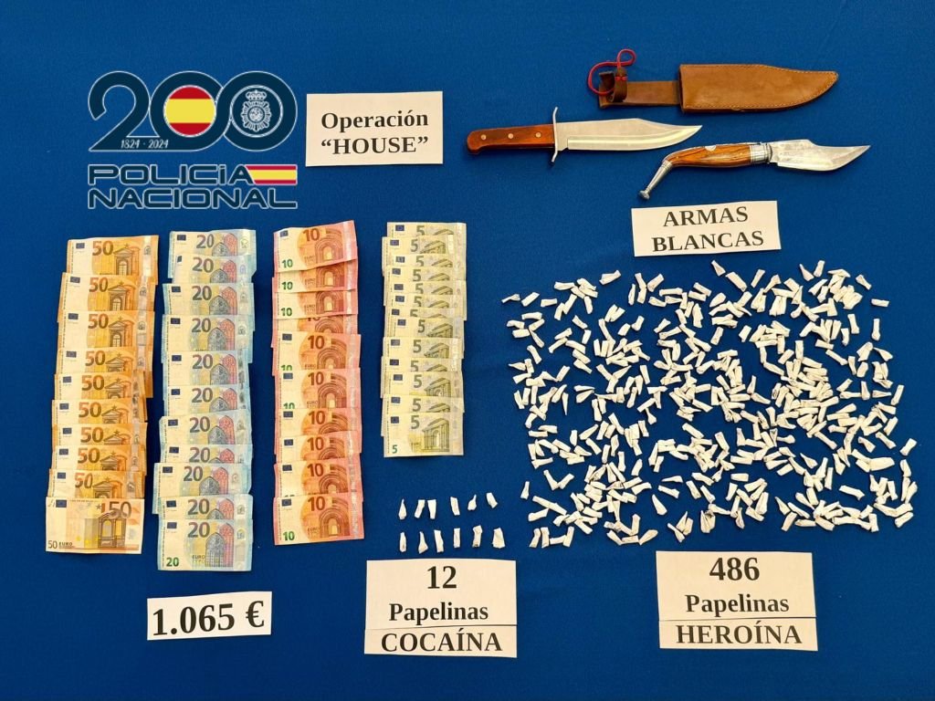 Material incautado por la Policía Nacional en Jerez en la Operación House contra el tráfico de drogas, mostrando billetes ordenados de diverso valor que suman 1.065 euros, armas blancas consistentes en un cuchillo de caza y una navaja grande, 12 papelinas de cocaína y 486 papelinas de heroína extendidas sobre una superficie azul junto al logotipo del 200 aniversario policial.