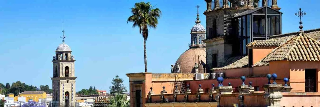 Panorámica de los tejados del Alcázar de Jerez de la Frontera bajo un cielo azul, mostrando la torre del campanario de la catedral, cúpulas barrocas, una palmera alta y detalles arquitectónicos históricos de piedra y tonos rojizos.