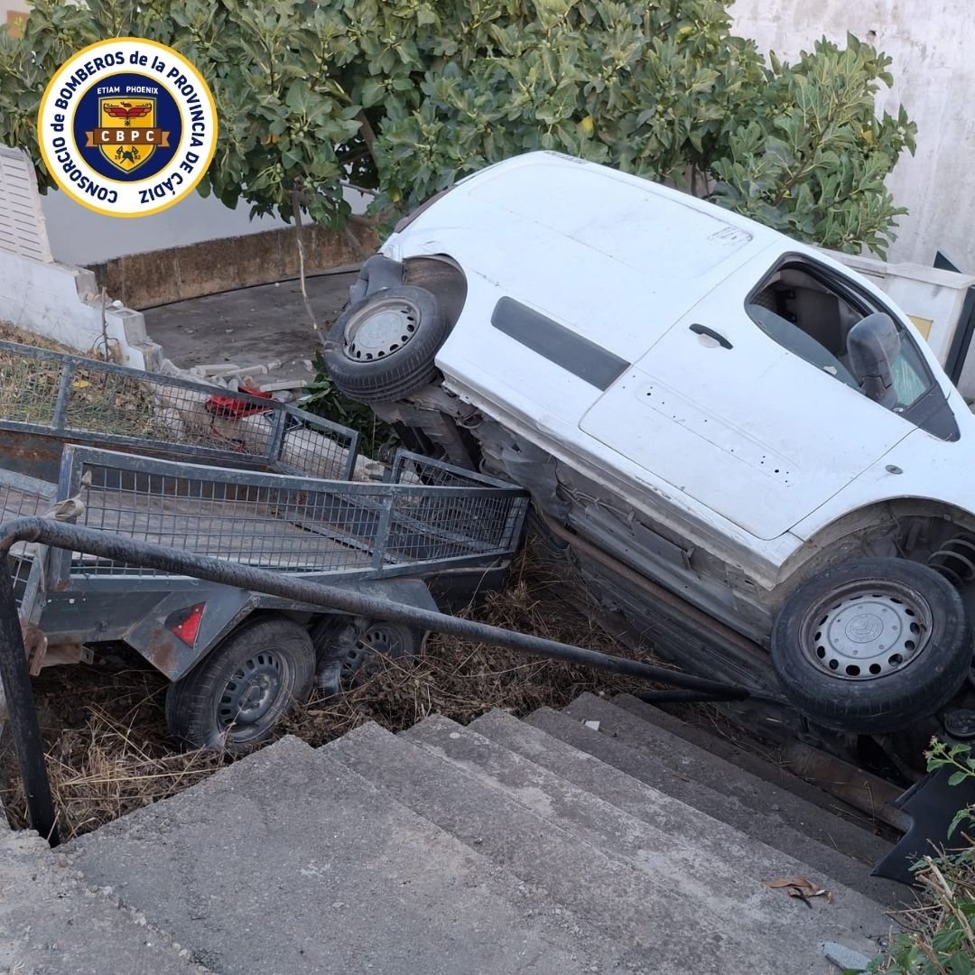 Furgoneta blanca con remolque accidentada y empotrada en posición vertical sobre unas escaleras de hormigón junto a un muro y vegetación, con el logotipo del Consorcio de Bomberos de la Provincia de Cádiz.