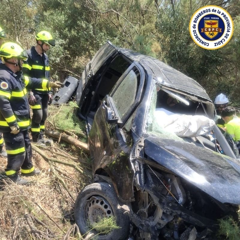 Efectivos de bomberos de Jerez inspeccionan un vehículo negro gravemente dañado y volcado en un terraplén arbolado durante las tareas de excarcelación en la autovía A382.