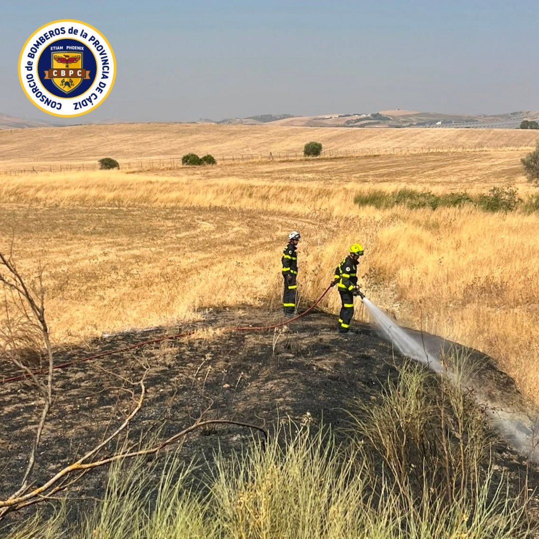 Dos bomberos extinguiendo un incendio de pastos con una manguera en la cuneta de la carretera CA-5200.