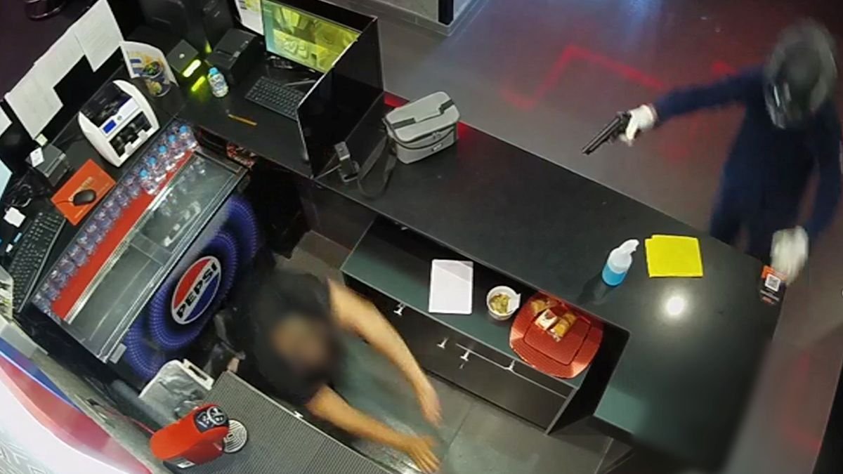 Captura de pantalla de la cámara de seguridad que muestra a uno de los asaltantes con casco y arma corta apuntando al cajero, quien se encuentra en el suelo detrás del mostrador del salón de juegos.