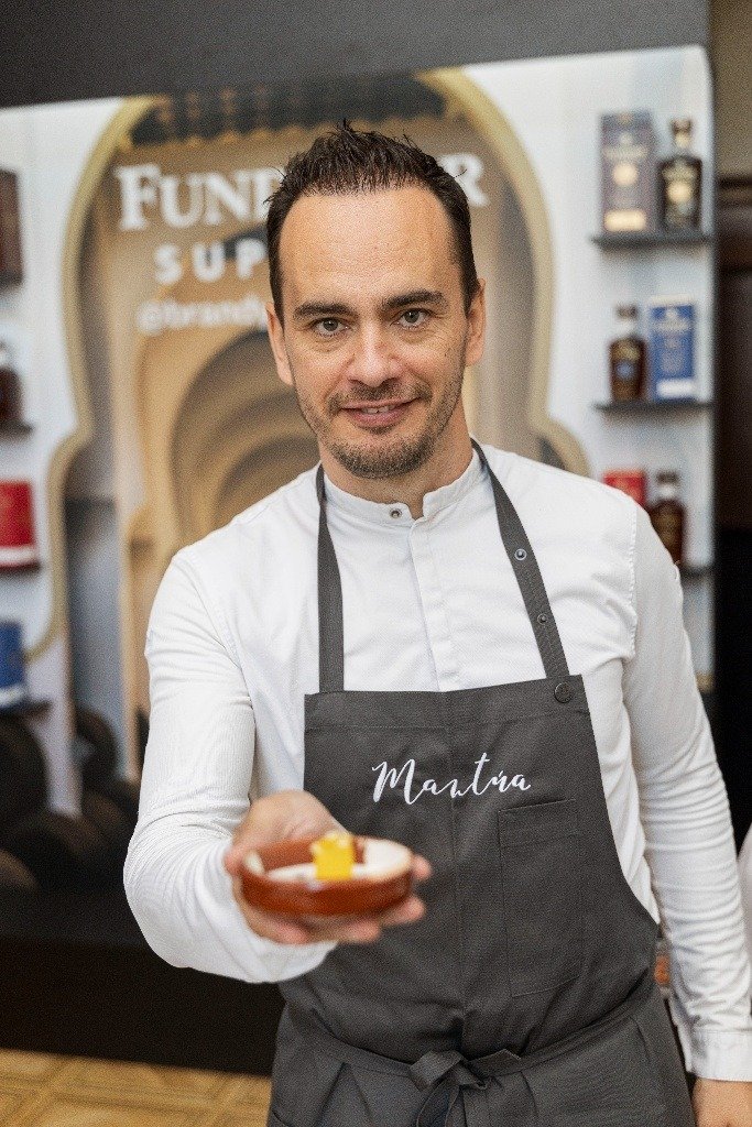 El chef jerezano Israel Ramos, con una camisa blanca y delantal gris con el nombre de su restaurante Mantra, ofrece en primer plano una cazuela de barro pequeña con una tapa tradicional durante la presentación de Jerez Capital Española de la Gastronomía.