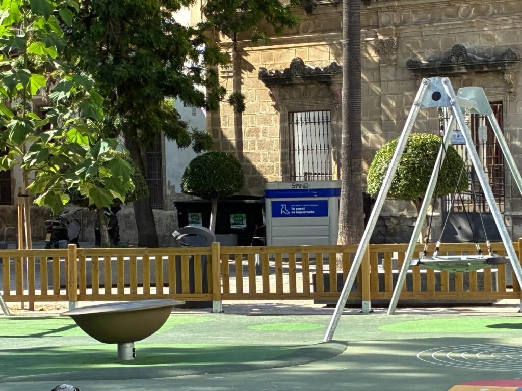 Contenedor de papel instalado junto a la valla de un parque infantil en la Plaza del Progreso, en el centro histórico de Jerez.