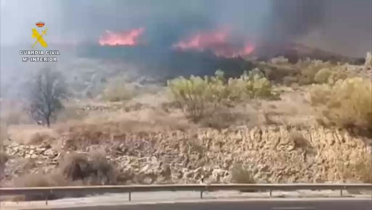Vista desde una carretera donde se observa un incendio forestal activo quemando vegetación en una colina cercana bajo el logo de la Guardia Civil.