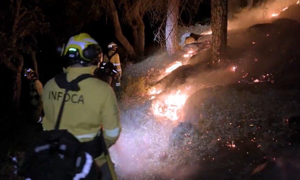 Bombero forestal del Plan Infoca de espaldas con chaqueta amarilla, casco y mochila, observando focos de fuego activos en una ladera boscosa durante la noche.