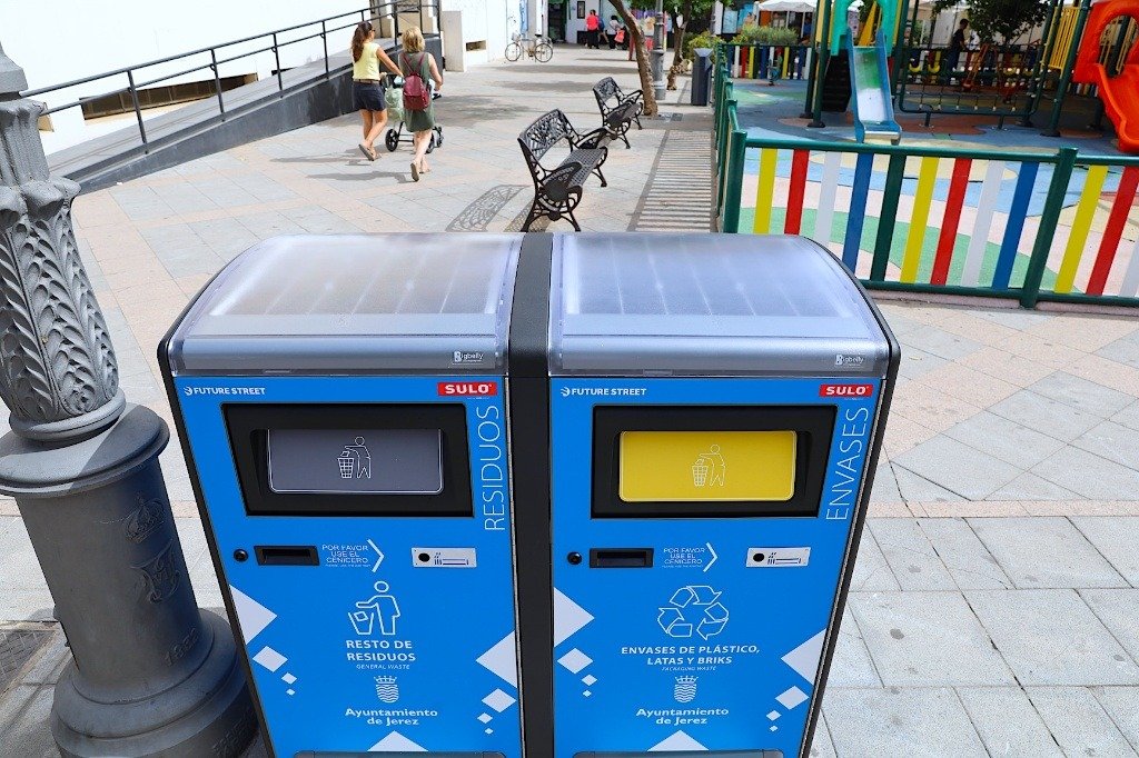 Primer plano de dos nuevas papeleras compactadoras solares azules de Sulo Ibérica instaladas en una plaza de Jerez, una para residuos generales y otra para envases, con sus paneles solares visibles en la parte superior y logotipos municipales.