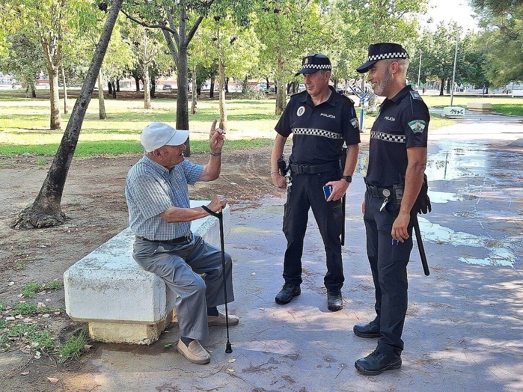 Dos agentes de la Policía Local conversan amablemente con un hombre mayor sentado en un banco de un parque, reflejando una labor de atención ciudadana y asistencia humanitaria.