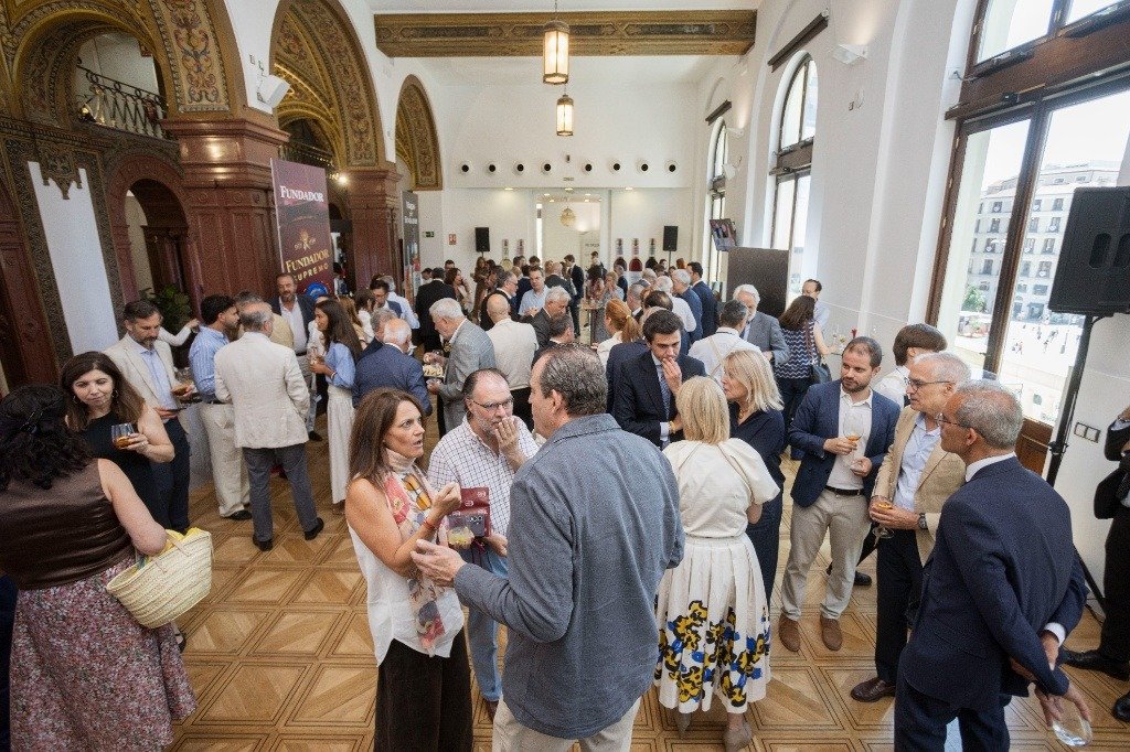 Vista amplia de un salón señorial con ventanales altos donde se congregan decenas de invitados en corrillos, conversando y disfrutando de un aperitivo con copas de vino durante la presentación oficial de Jerez Capital Española de la Gastronomía 2026. Al fondo, se aprecia un cartel publicitario de las bodegas Fundador.