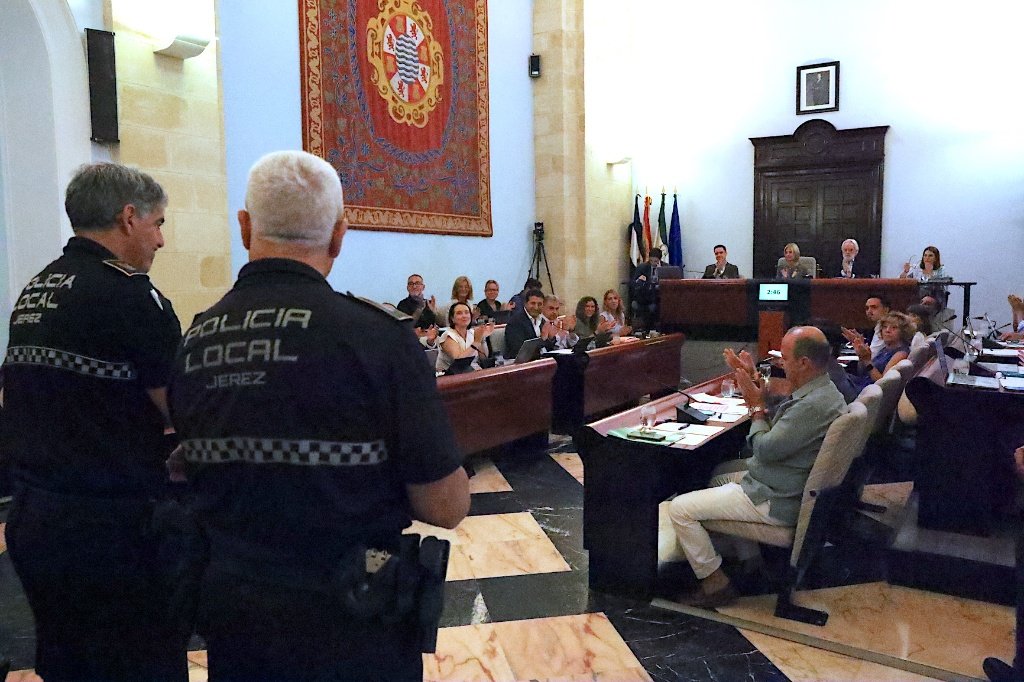 Un plano general del Salón de Plenos de Jerez, donde los concejales y la presidencia aplauden. En primer plano, de espaldas, hay dos agentes uniformados de la Policía Local de Jerez con el distintivo de la ciudad en la espalda.