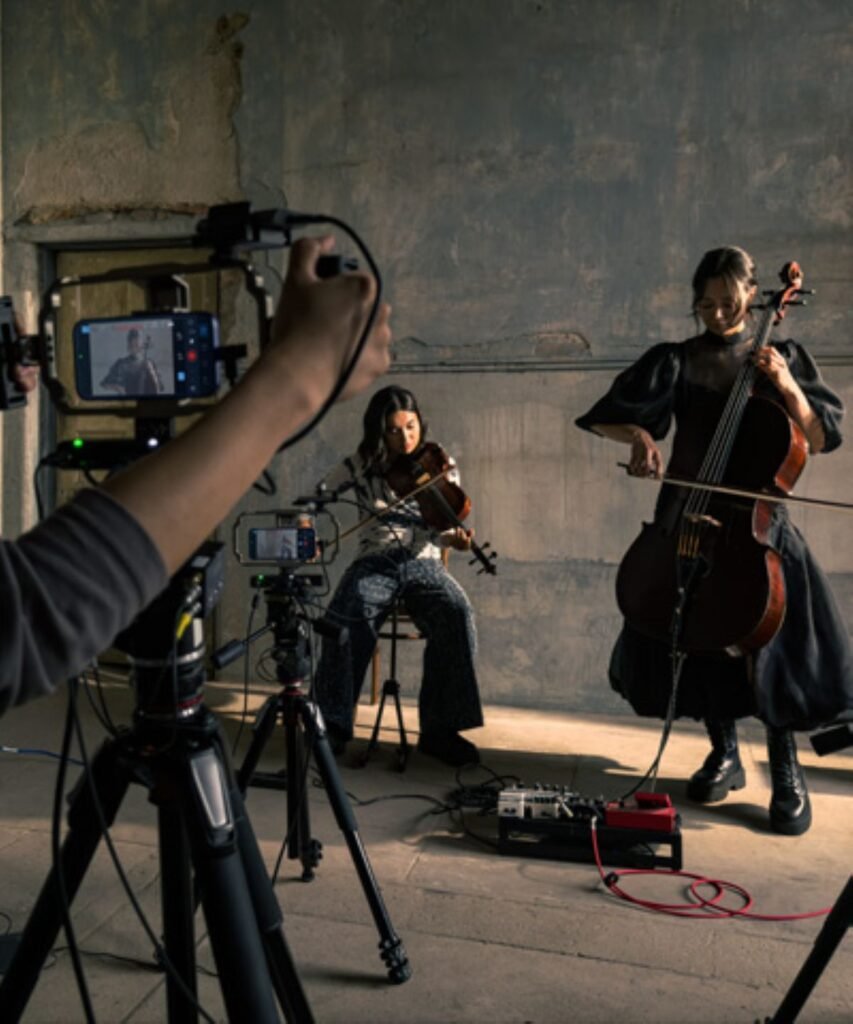 Una toma de detrás de escena en un entorno rústico y texturizado, que muestra a dos músicas clásicas (una violinista sentada y una violonchelista de pie) actuando para un equipo de rodaje. El primer plano está dominado por un complejo sistema de cámara de iPhone 17 Pro, donde una mano sujeta un estabilizador que integra un iPhone 17 Pro Genlock synchronized camera controller, con el texto 'iPhone 17 Pro Genlock' claramente visible en el dispositivo y la interfaz de usuario de sincronización en la pantalla. Múltiples trípodes de cámara, cada uno con controladores Genlock y iPhones 17 Pro, están dispuestos alrededor de los músicos. La violinista está sentada a la izquierda y la violonchelista está de pie a la derecha, ambas vestidas de negro y concentradas en su interpretación. El suelo de hormigón está cubierto de cables profesionales y un tablero de pedales de efectos. La luz del sol entra por una abertura, iluminando a los músicos.