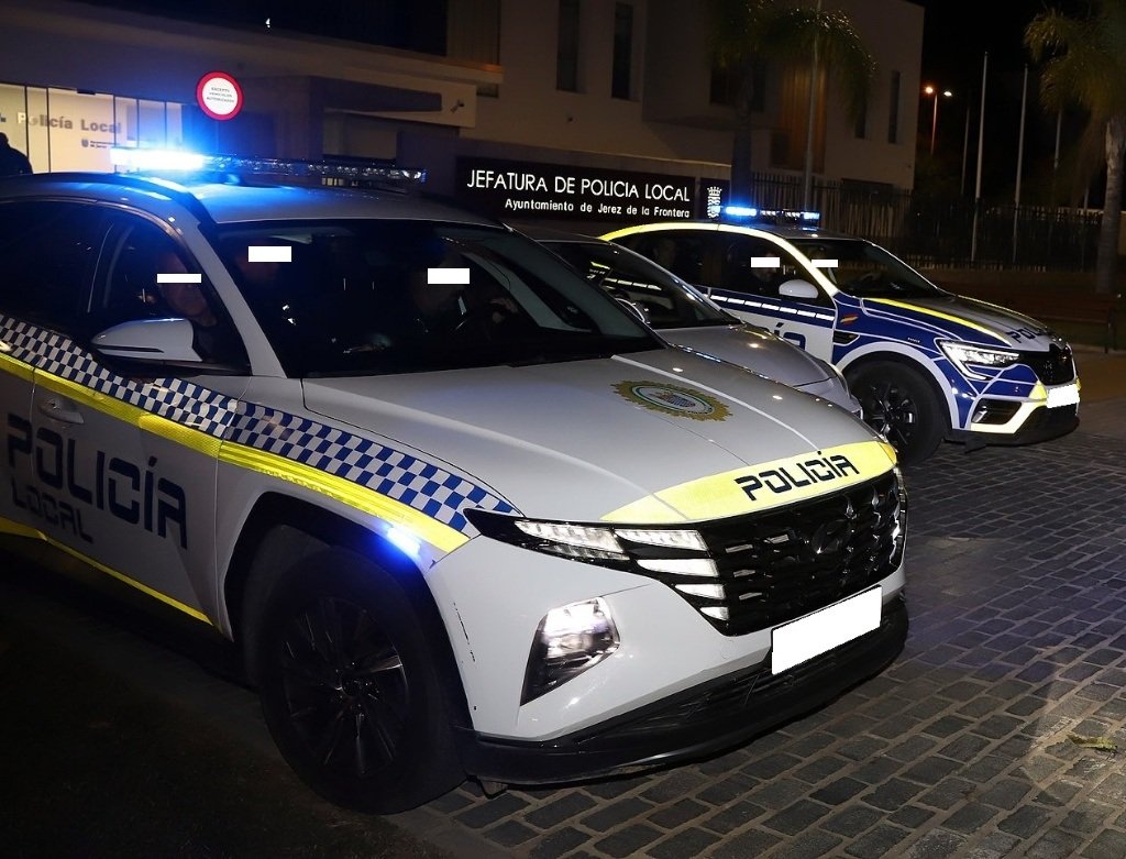 Coches patrulla de la Policía Local de Jerez estacionados frente a la Jefatura con las luces de emergencia encendidas durante la noche.