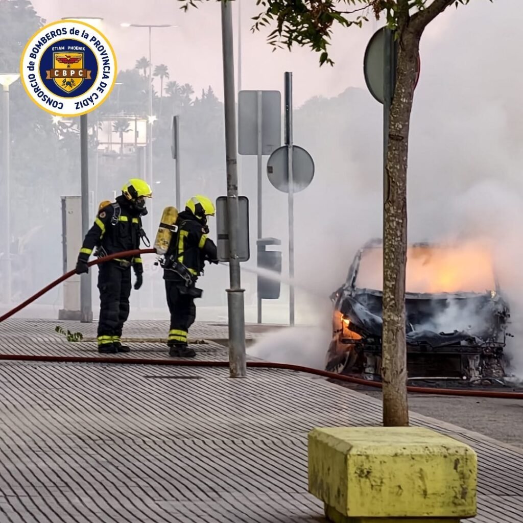 Dos bomberos equipados con trajes de protección y manguera sofocan el incendio de un taxi en la avenida Juan Carlos I de Jerez de la Frontera.