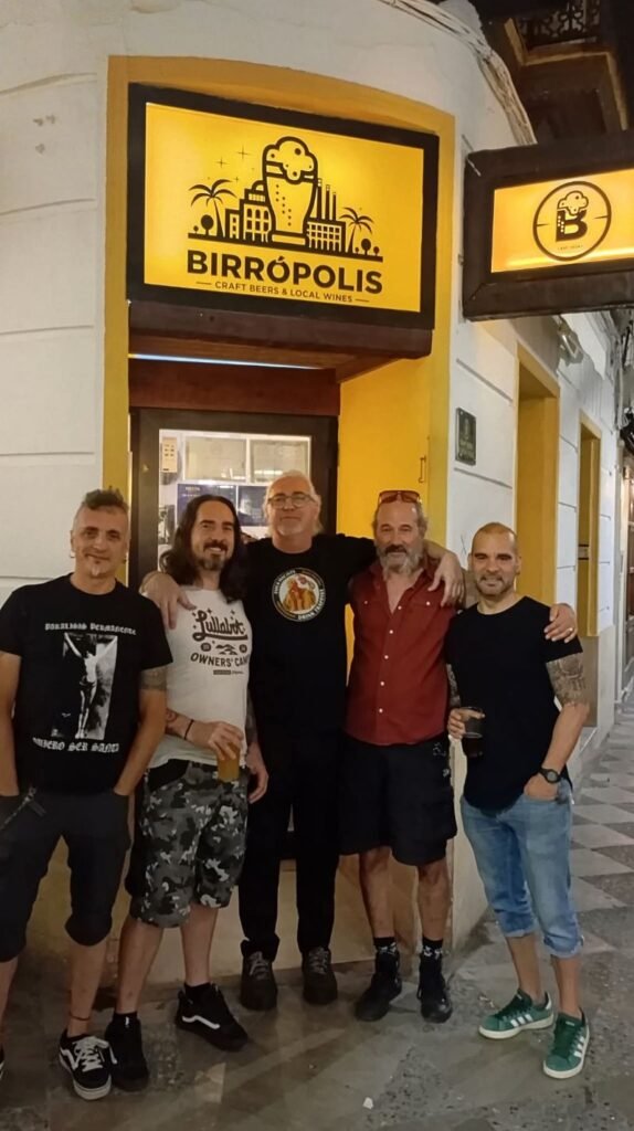 Los integrantes del grupo de rock jerezano Hades posan sonriendo junto a Jimmy en la entrada de la cervecería Birrópolis en Jerez, bajo los carteles iluminados.