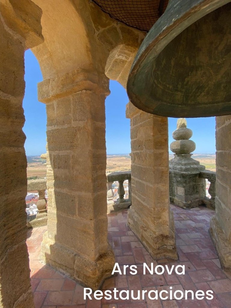 Perspectiva desde el interior del campanario de Santa María la Coronada, mostrando los pilares de piedra restaurados, el suelo de baldosas de barro, una gran campana en primer plano y la balaustrada con vistas al horizonte de Medina Sidonia.