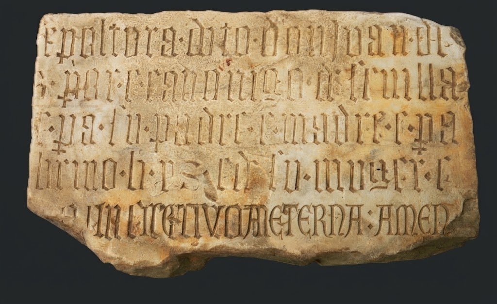 Vista detallada y frontal de la lápida funeraria gótica de piedra caliza de 1478, con su inscripción latina en minúsculas góticas, contra un fondo de museo oscuro.