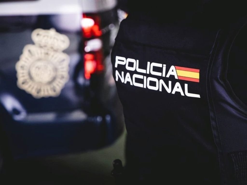 Un primer plano enfocado en la parte posterior del chaleco táctico de un agente de Policía Nacional de España. El texto blanco "POLICIA NACIONAL" está claramente visible junto a una pequeña bandera de España. Al fondo, fuera de foco, se aprecia un coche patrulla con su emblema y luces traseras rojas.