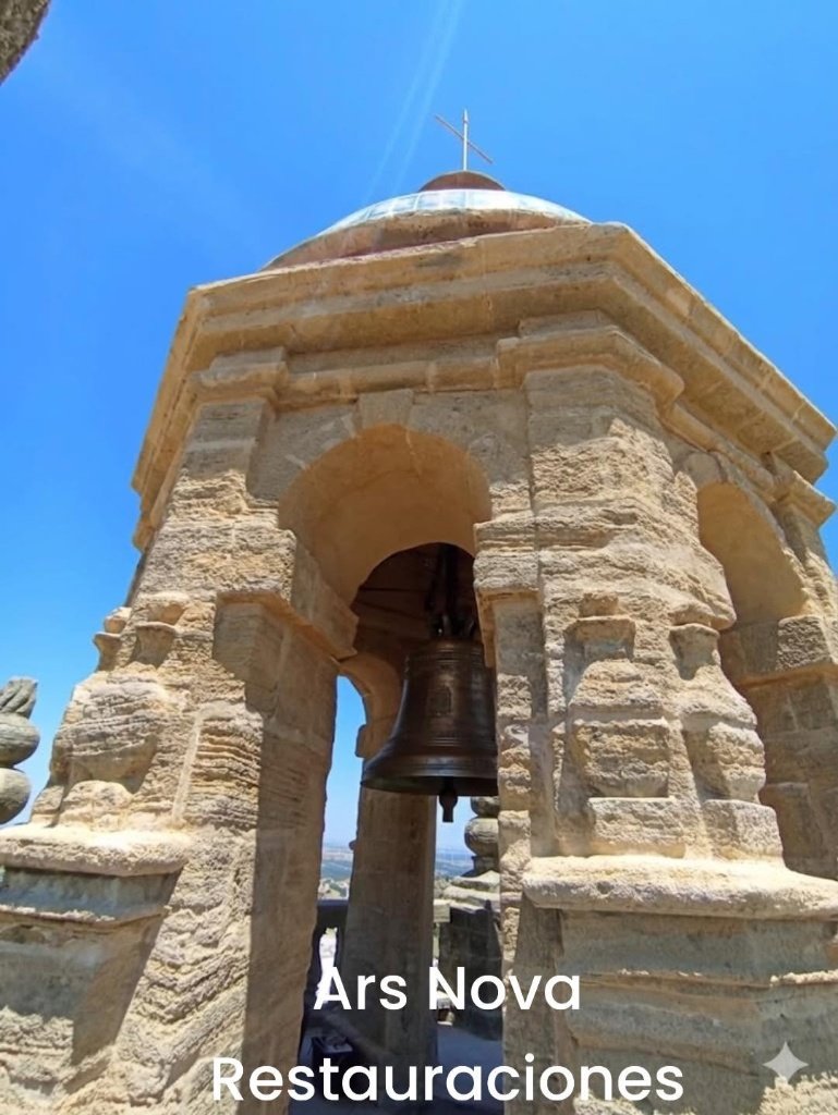 Plano contrapicado del templete octogonal de la torre de Santa María la Coronada en Medina Sidonia, mostrando los arcos de piedra arenisca limpia, una campana en el centro y la cúpula con la cruz superior bajo un cielo azul intenso.