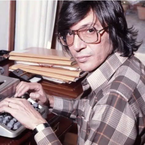 Fotografía de primer plano de Manuel Alejandro, joven y con gafas, sentado ante una máquina de escribir, con un fajo de partituras sobre el mueble y una expresión concentrada.