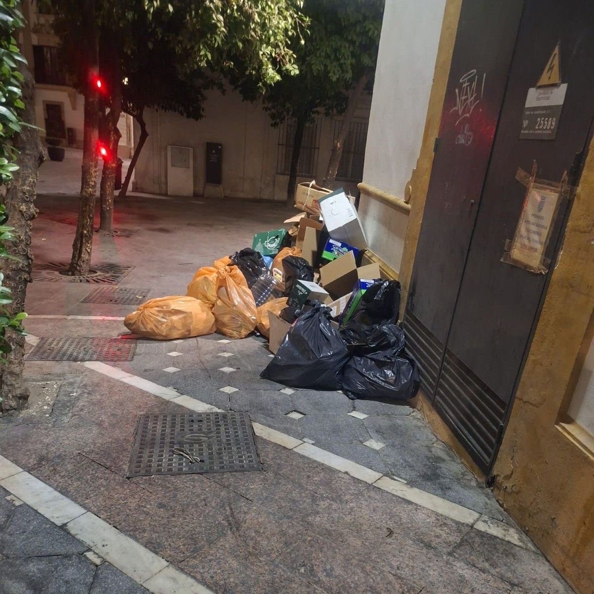 Perspectiva nocturna de la esquina de la calle Belén con bolsas de basura naranjas y negras apiladas junto a una fachada en el centro de Jerez.