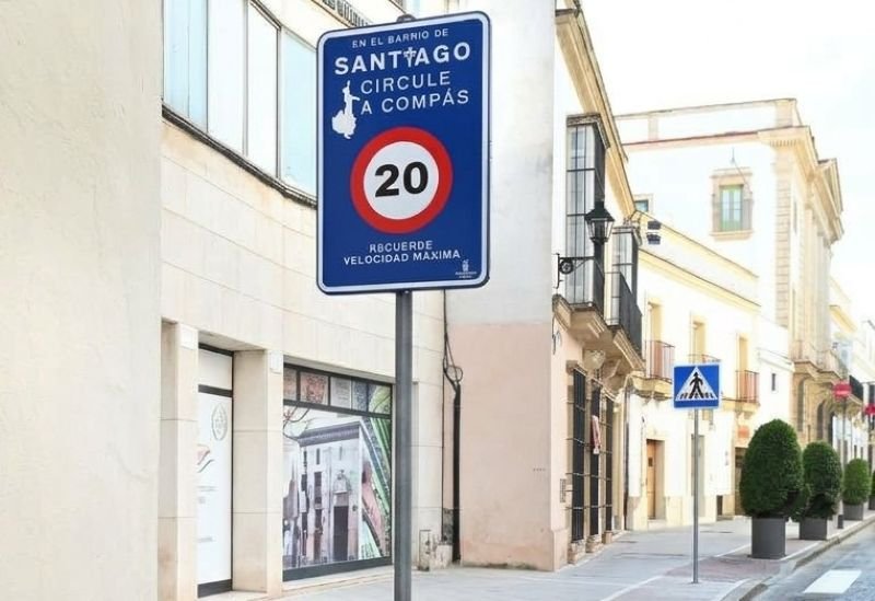 Fotografía a pie de calle en Jerez de la Frontera, mostrando una señal de tráfico azul no homologada en el Barrio de Santiago que reza: 'EN EL BARRIO DE SANTIAGO CIRCULE A COMPÁS' y establece un límite de '20' km/h con el recordatorio 'RECUERDE VELOCIDAD MÁXIMA'. La señal está situada frente a edificios residenciales y comerciales de estilo tradicional, con la calle adoquinada y la marca jerezsinfronteras.es en la esquina inferior izquierda.