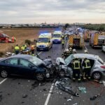 Fotografía aérea de un grave accidente de tráfico en una carretera de dos carriles, donde dos turismos han chocado frontalmente en el kilómetro 1. Múltiples equipos de emergencia están presentes: un helicóptero médico está aterrizando en un campo adyacente, varias ambulancias con luces azules y coches de la Guardia Civil están estacionados en la carretera. Bomberos con uniformes reflectantes están trabajando para liberar a una persona atrapada en uno de los vehículos accidentados. La escena es de alta tensión y despliegue de rescate en un entorno rural de campos de olivos.