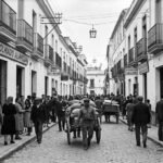 Fotografía antigua en blanco y negro de una concurrida calle peatonal en Jerez de la Frontera alrededor de 1930. En primer plano, varios peatones con vestimenta de la época caminan por el centro y las aceras, destacando un hombre empujando un carro de mano de dos ruedas. Los edificios a ambos lados de la calle tienen balcones con barandillas de hierro, farolas colgantes centrales y locales comerciales en las plantas bajas.